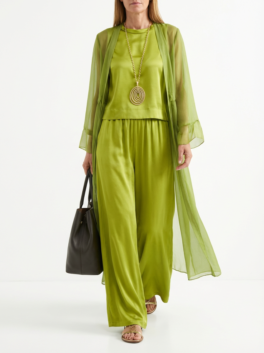 Model in an elegant three-piece olive green set with sheer duster, tank top, and wide-leg pants, accessorized with gold necklace and holding black bag in studio setting.