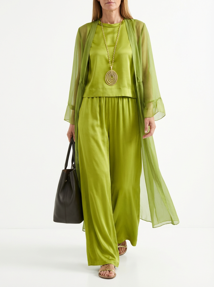 Model in an elegant three-piece olive green set with sheer duster, tank top, and wide-leg pants, accessorized with gold necklace and holding black bag in studio setting.