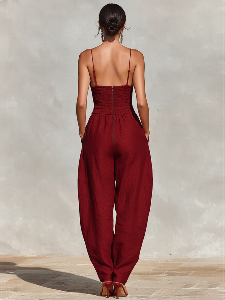 Back view of woman in deep red jumpsuit, showing spaghetti straps and shirred back, on stone surface.