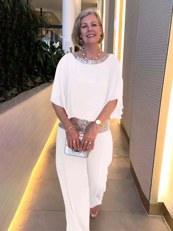 Woman in a white elegant set with shimmering belt and neckline, holding a silver clutch. Indoor setting with plants.