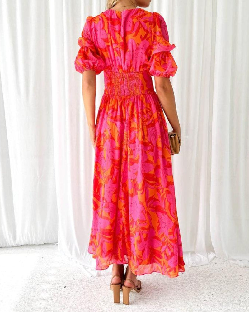 Back view of a flowing long dress in pink and orange floral pattern, highlighting the smocked waist and ruffled sleeves. White curtain background.