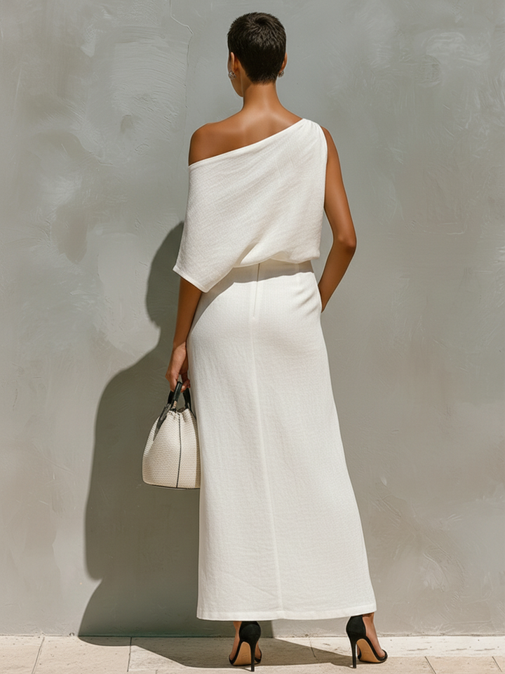 Back view of a model in a white off-shoulder dress holding a textured handbag, standing against a neutral outdoor wall.