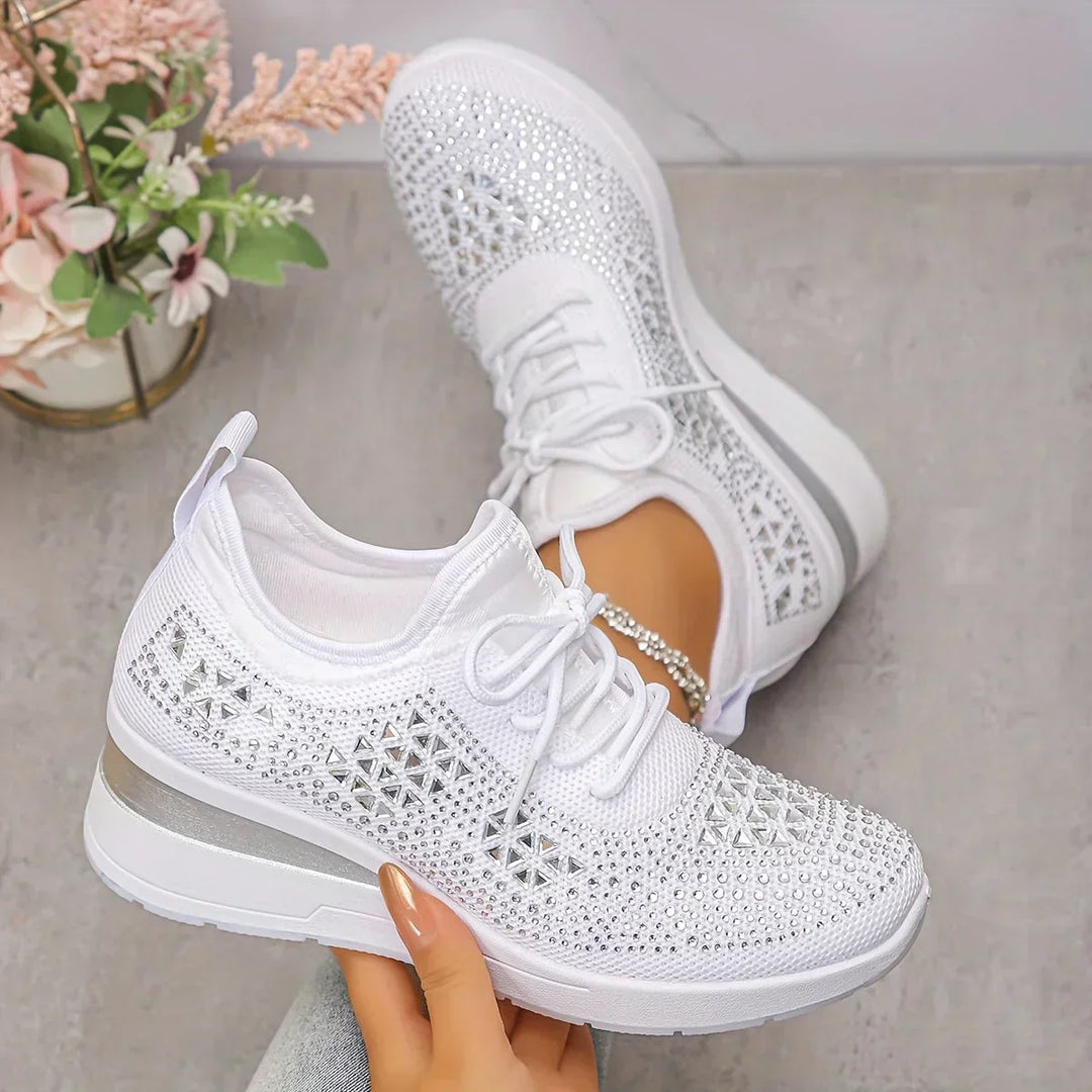 White orthopedic sneakers with glimmering accents held in hand, set on a light concrete surface with flowers nearby.