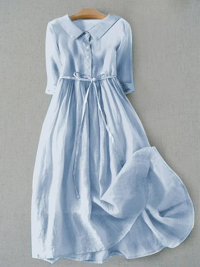 Light blue short sleeve dress with buttons and tie waist, hanging on a wooden hanger against a gray background.