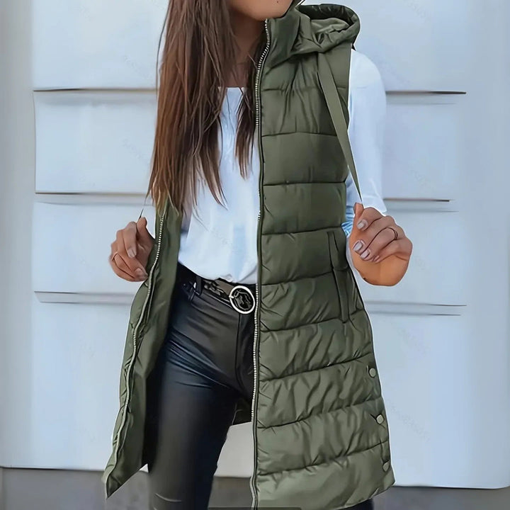 Dark green sleeveless puffer vest with hood worn over white top and black pants, outdoor setting.