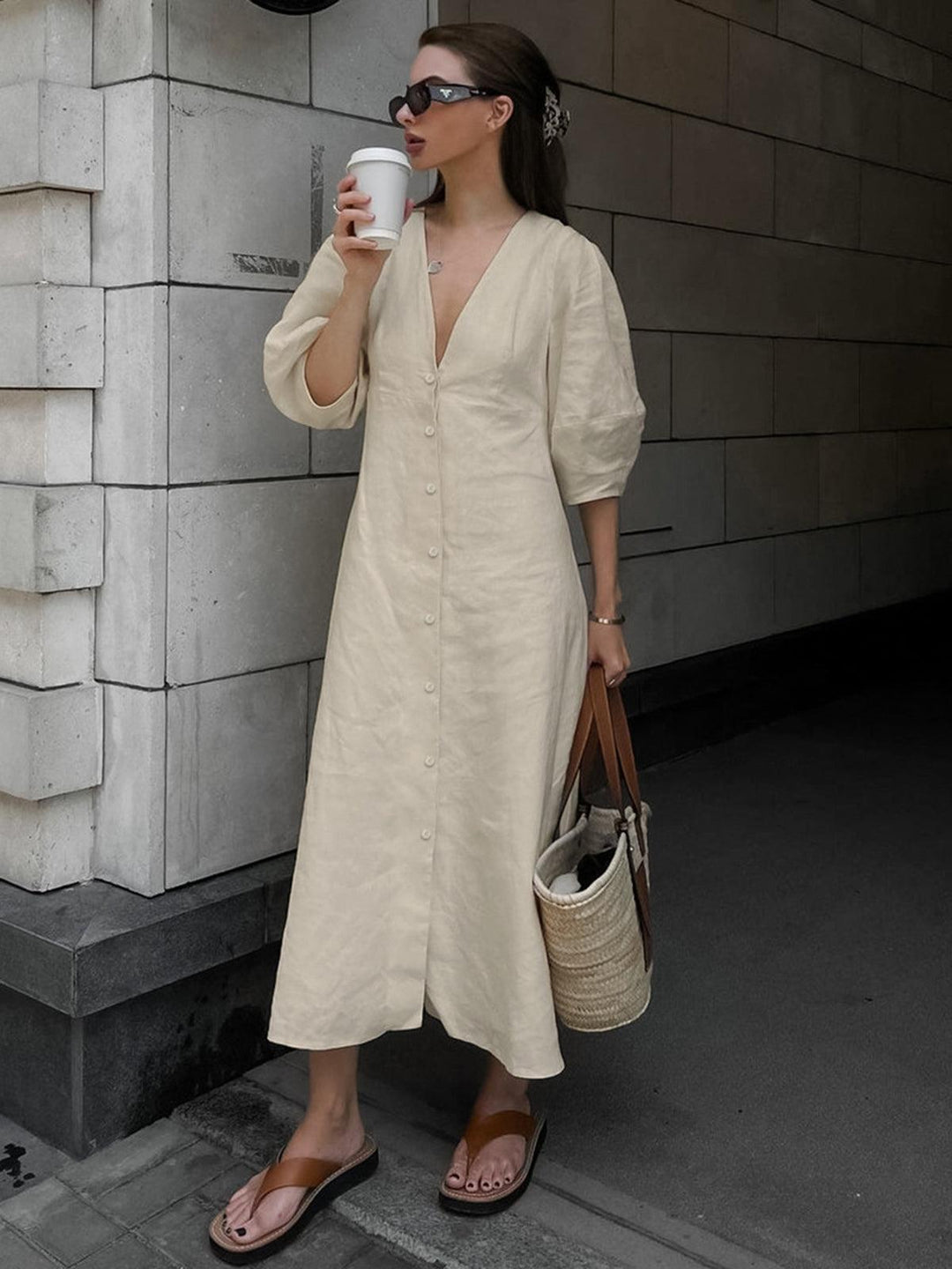 Woman in cream buttoned maxi dress holding coffee in urban setting, wearing sunglasses and sandals, with tote bag.