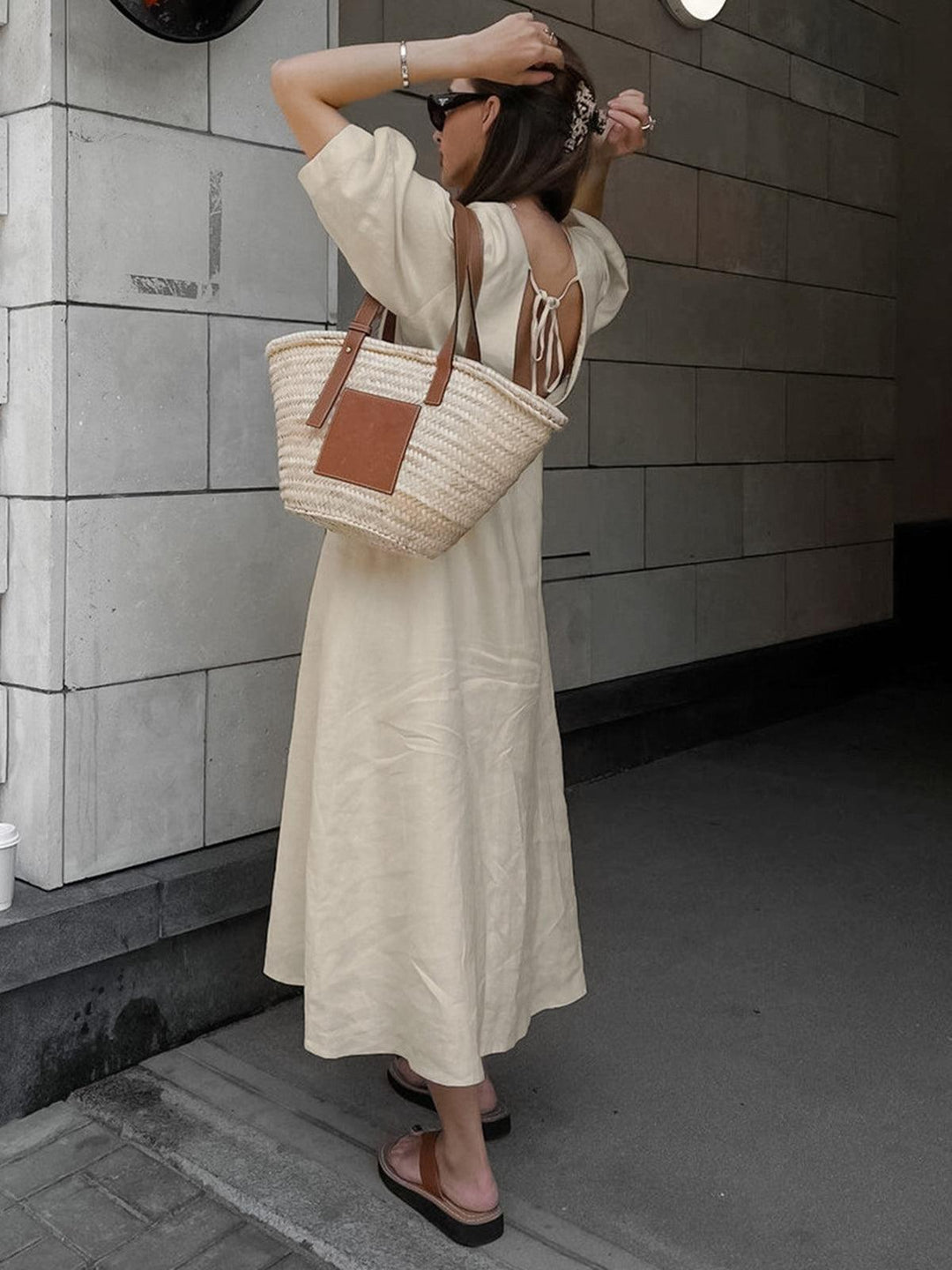 Back view of woman in cream buttoned maxi dress adjusting hair in urban setting, with woven tote bag.