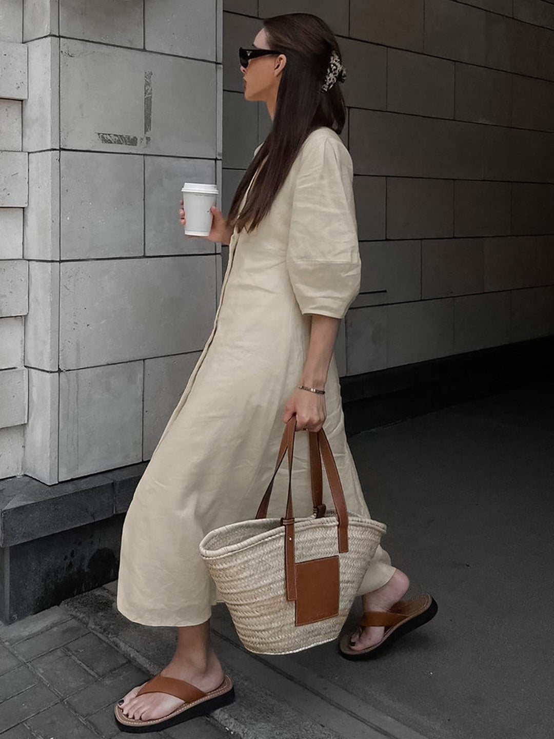 Side view of woman in cream buttoned maxi dress holding coffee in urban setting, wearing sunglasses, with tote bag.