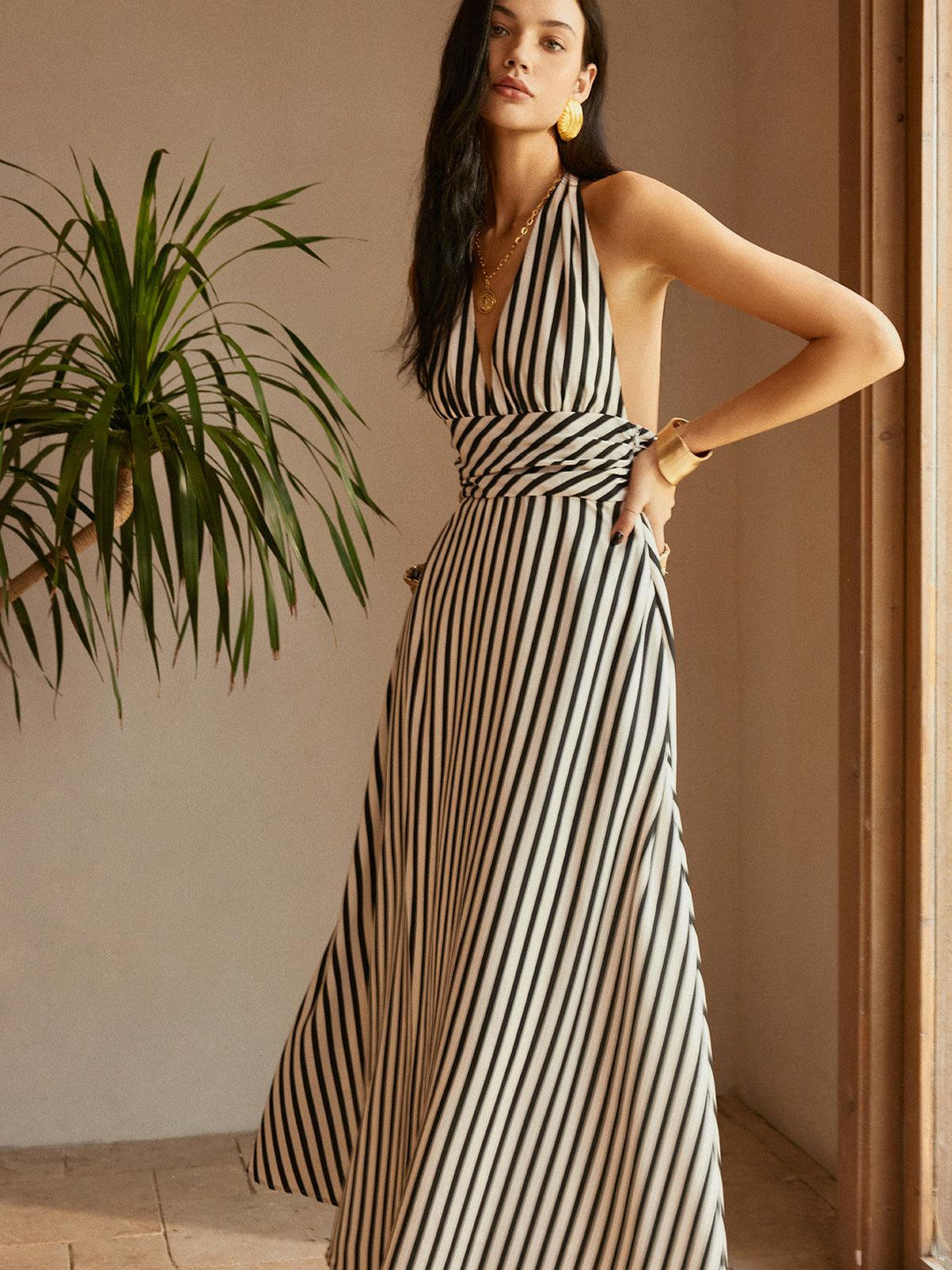 Model wearing backless striped dress indoors, posing with hand on hip, near plant and wooden door.