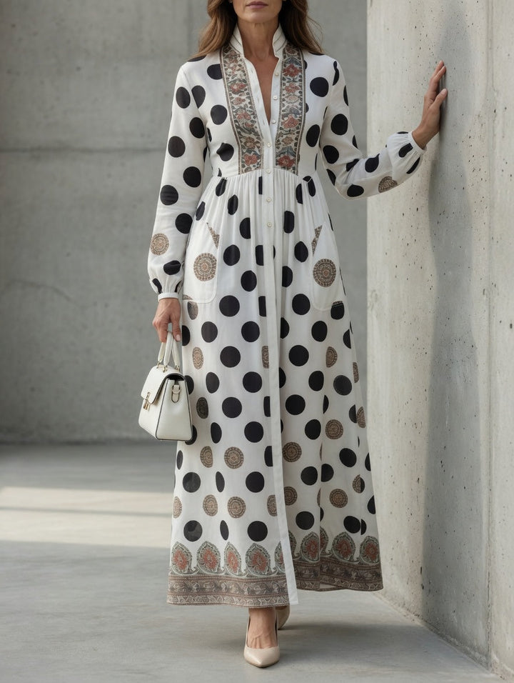 Elegant maxi dress adorned with polka dots and ornate designs, styled with a white handbag and heels against a concrete wall.
