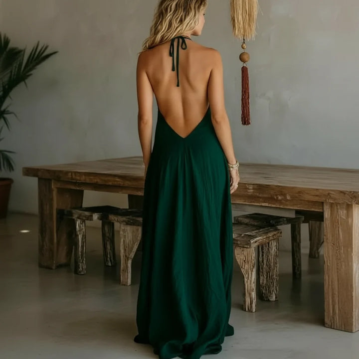 Back view of a woman wearing a green halter maxi dress in a rustic room with a wooden table and wall decor.