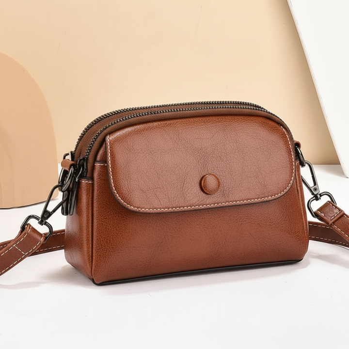 Brown crossbody bag with front pocket and adjustable strap, set on a beige surface with abstract shapes in the background.