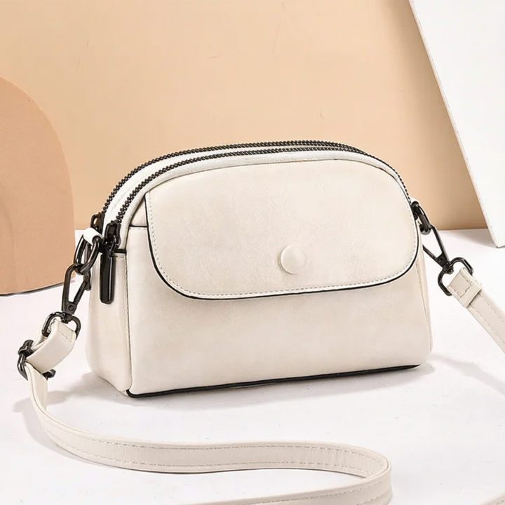 White crossbody bag showing a front pocket and adjustable strap, set on a beige surface with decorative shapes behind.