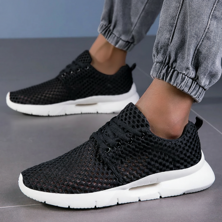Black orthopedic sneakers with mesh design worn with gray joggers on tiled floor.