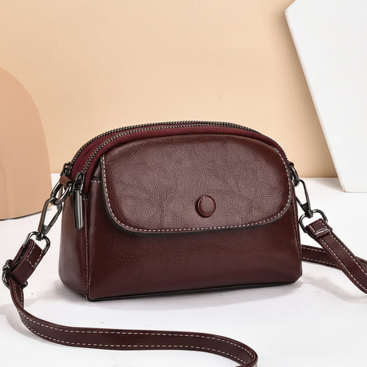 Maroon crossbody bag with front pocket and adjustable strap, placed against a beige surface with artistic shapes in the background.