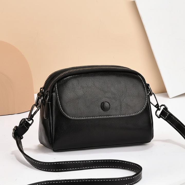 Black crossbody bag featuring a front pocket and adjustable strap, displayed on a beige backdrop with abstract shapes.