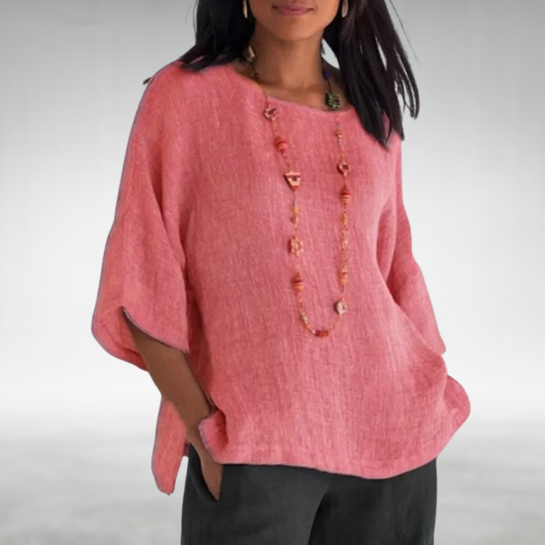 Woman wearing a pink boat-neck tunic with a long necklace, hands in pockets, against a neutral background.