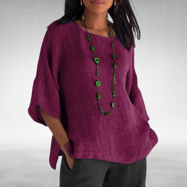 Woman in a purple boat-neck tunic with a green necklace, hands in pockets, gray backdrop.
