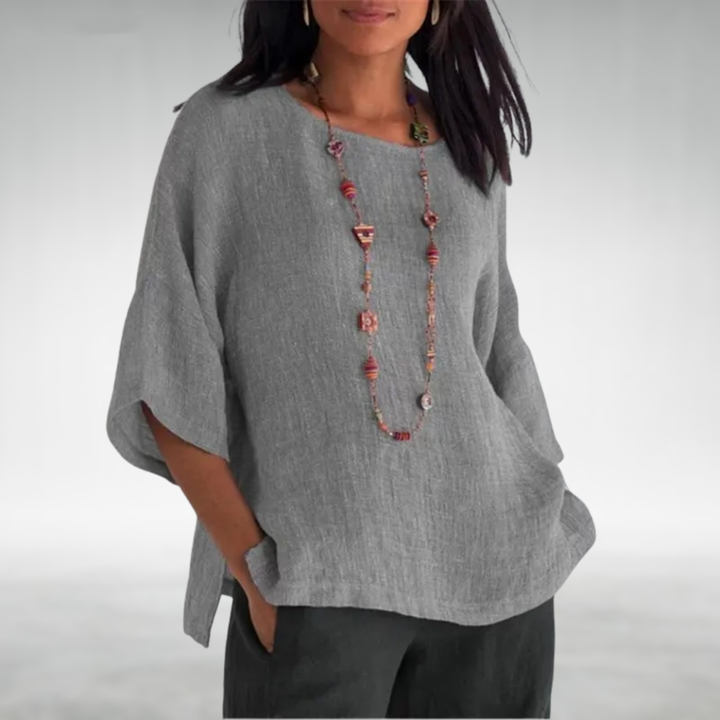 Woman in a gray boat-neck tunic with a colorful necklace, hands in pockets, light gray background.