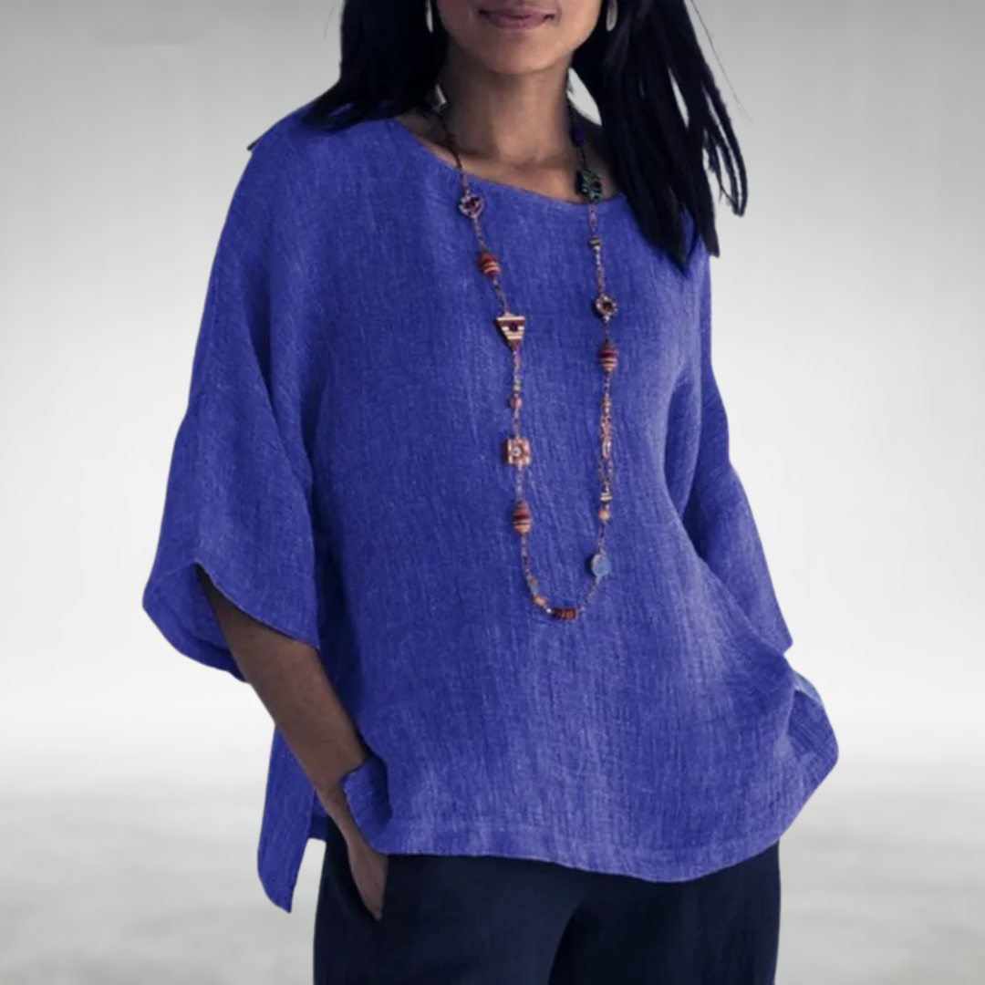 Woman wearing a blue boat-neck tunic with a long beaded necklace, hands in pockets, against a soft gray backdrop.