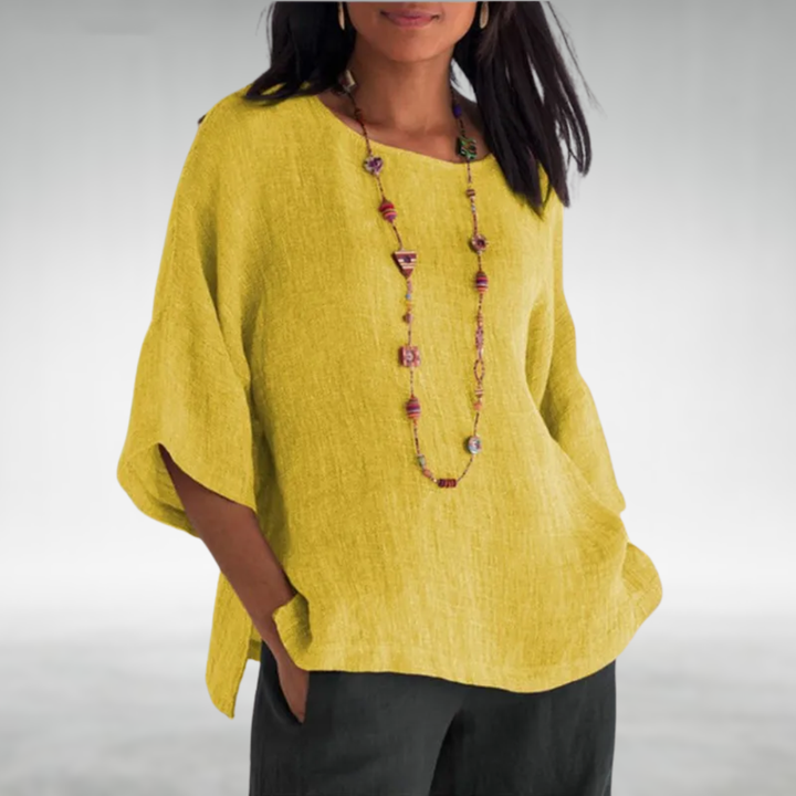 Woman in a yellow boat-neck tunic with a multi-colored necklace, hands in pockets, neutral background.