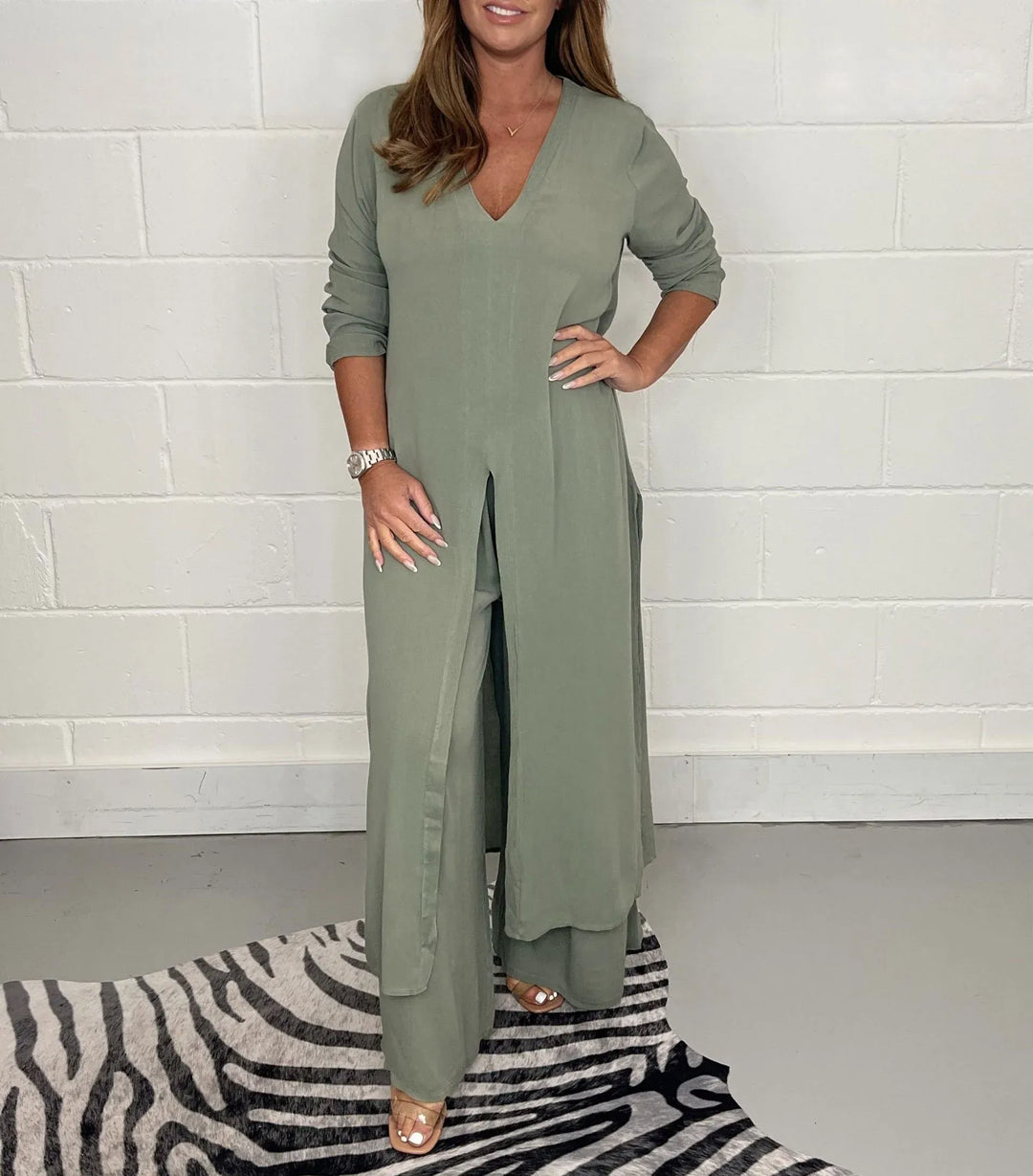 Model wearing a sage green two-piece set with long sleeves and a V-neck, standing on a zebra-patterned rug against a white brick wall.