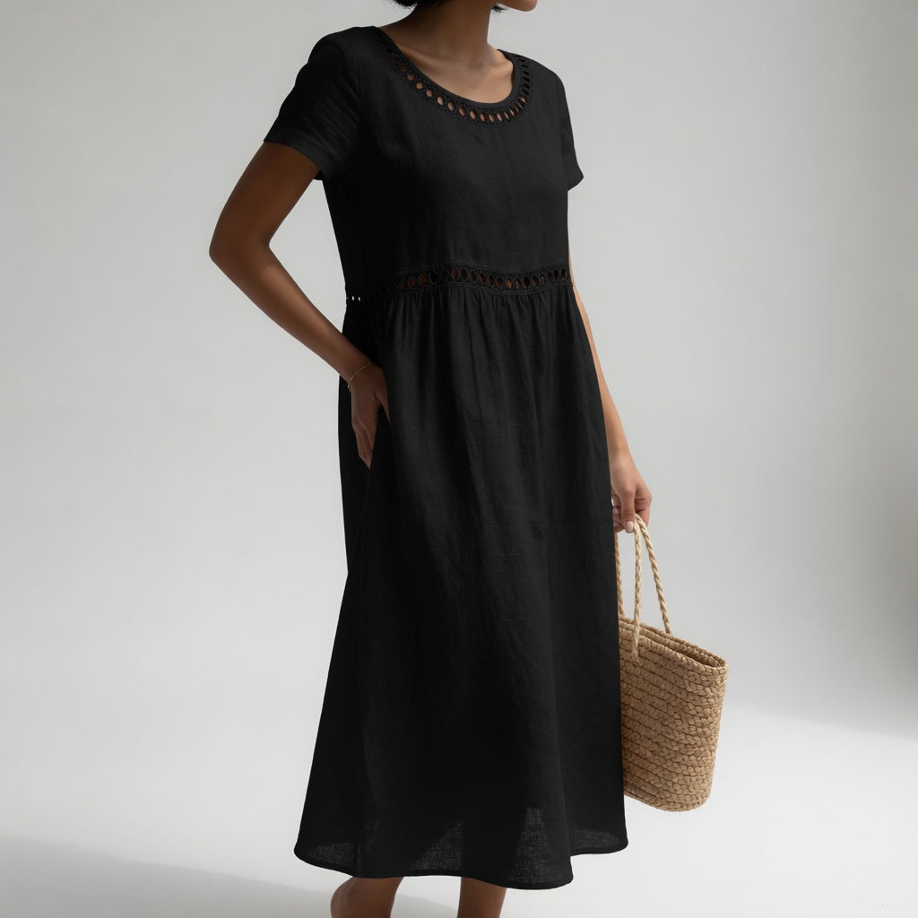 Black midi dress with eyelet trim, short sleeves, and a relaxed fit, held by a person with a woven bag. Studio light grey background.