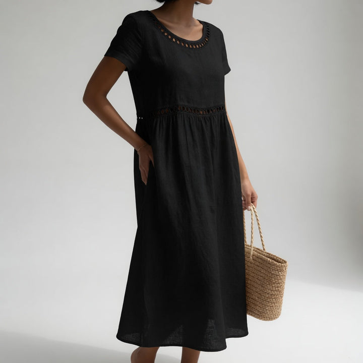 Black midi dress with eyelet trim, short sleeves, and a relaxed fit, held by a person with a woven bag. Studio light grey background.