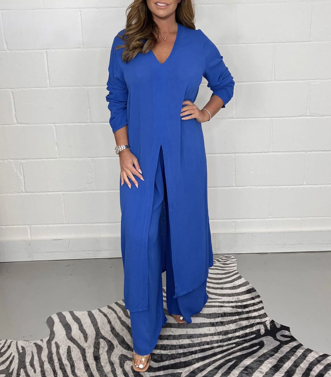 Model wearing a royal blue two-piece set with long sleeves and a V-neck, standing on a zebra-patterned rug against a white brick wall.