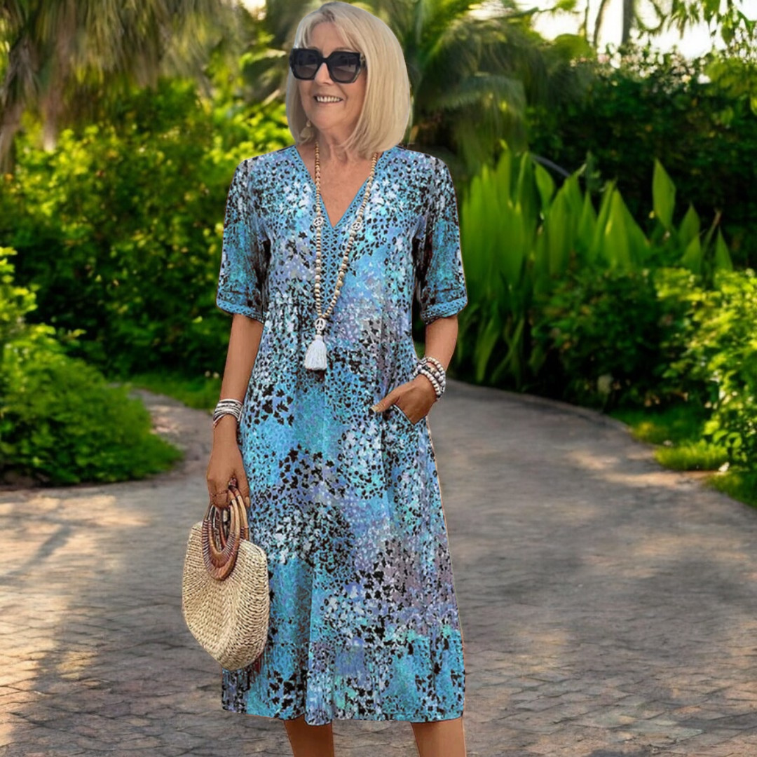 Woman in a blue and multicolored dress with abstract pattern, holding a straw bag on a garden path, surrounded by lush greenery.