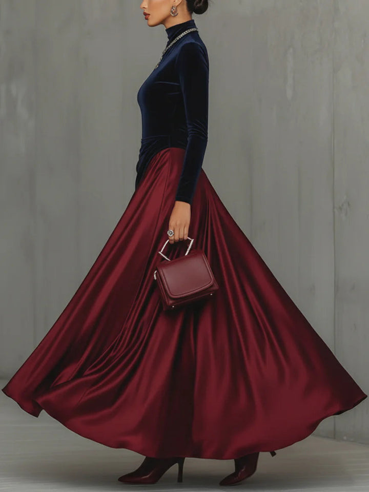 Model in a navy and maroon dress with a long flowing skirt and a handbag, side profile, set against a gray wall.