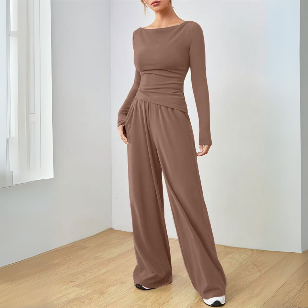 Model wearing brown long-sleeve top and wide-leg pants set in a minimalist indoor room with light walls and wooden floor.