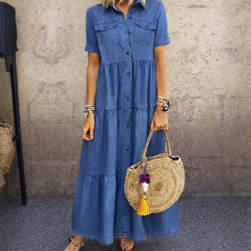 Dark blue button-front midi dress with short sleeves, tiered style, and front pockets. Accessorized with a round straw bag against a textured studio backdrop.