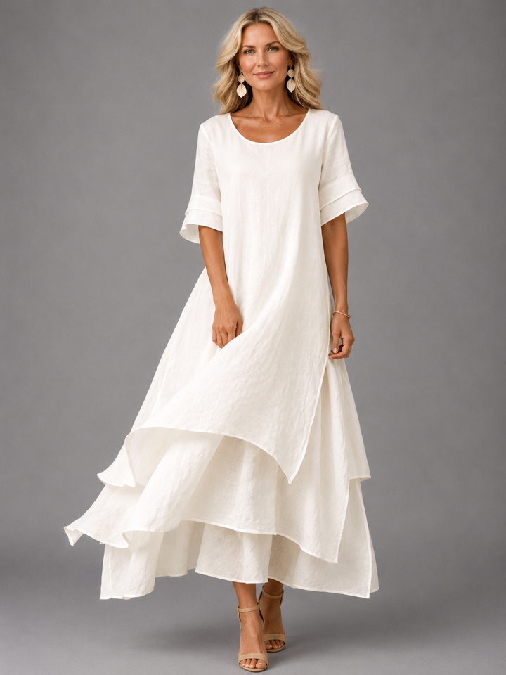 Woman in white flowing maxi dress with layered hem, posing in studio, smiling.