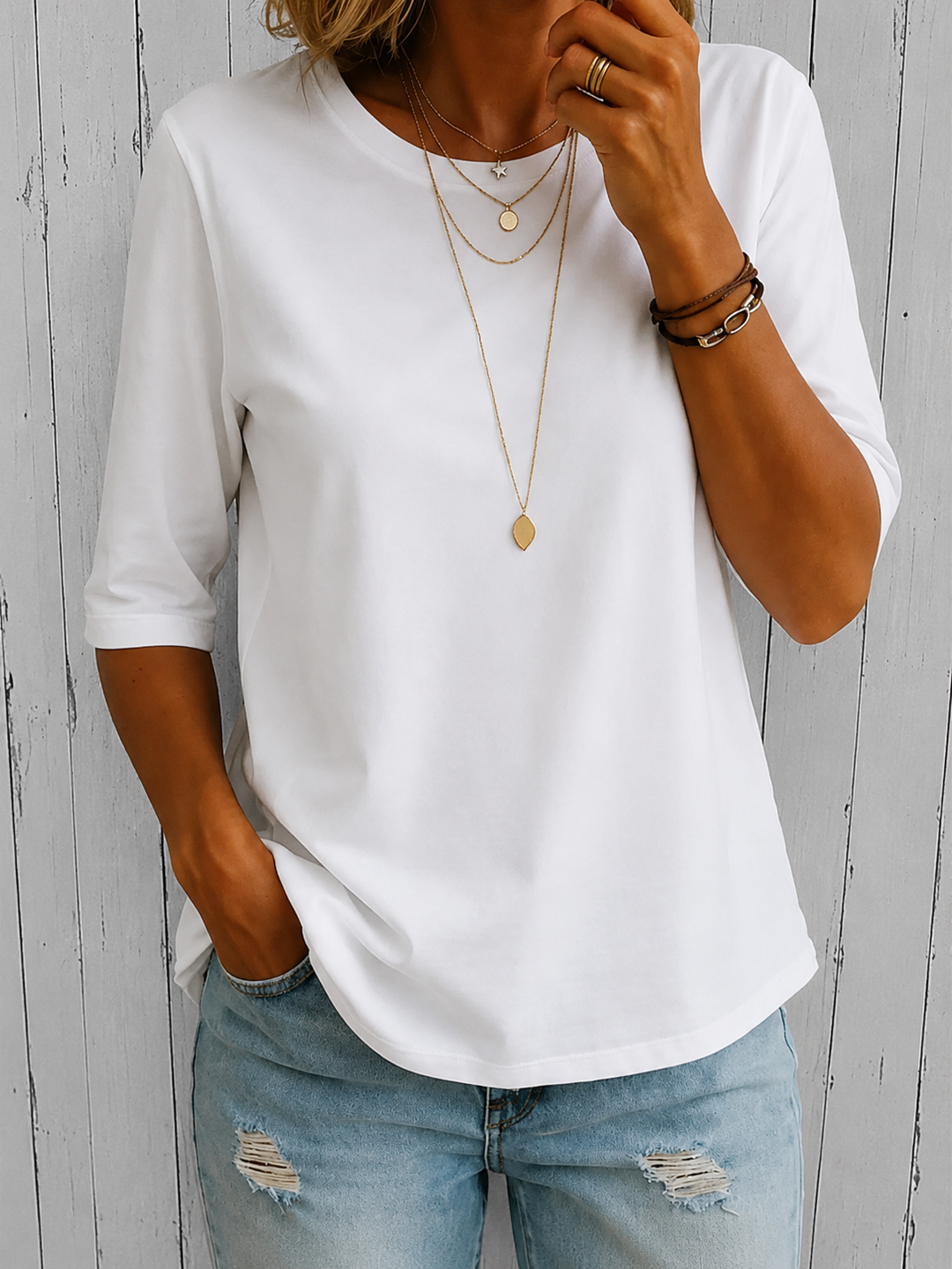 White three-quarter sleeve tee with layered necklaces, worn with light blue distressed jeans, against a rustic wood background.