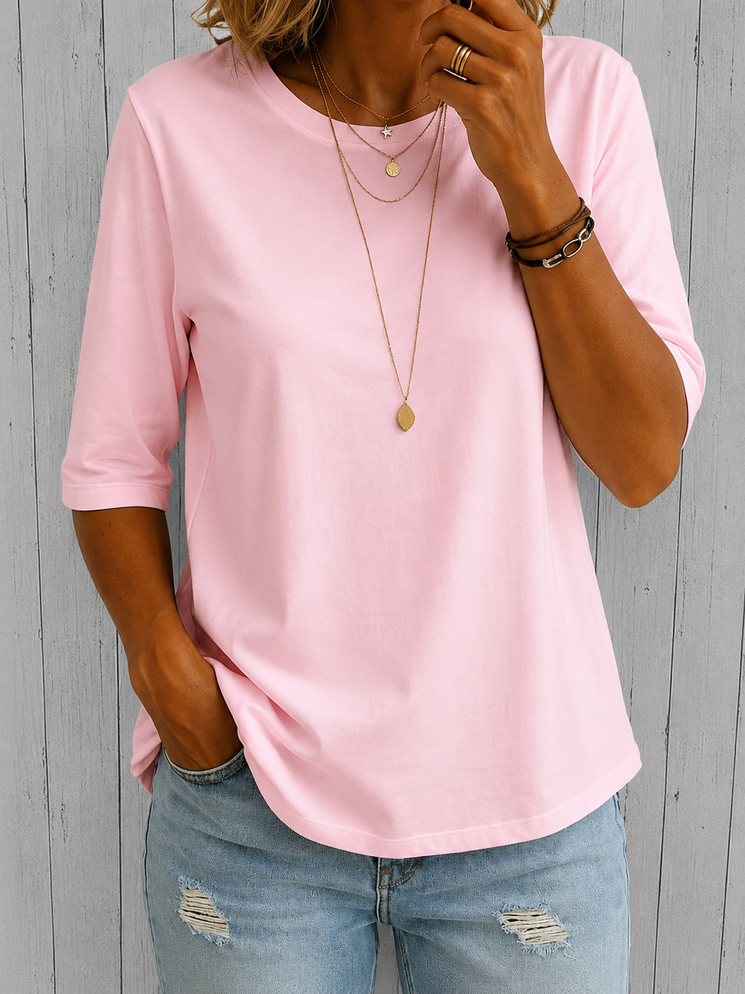 Pink three-quarter sleeve tee with layered necklaces, worn with light blue distressed jeans, against a textured panel background.