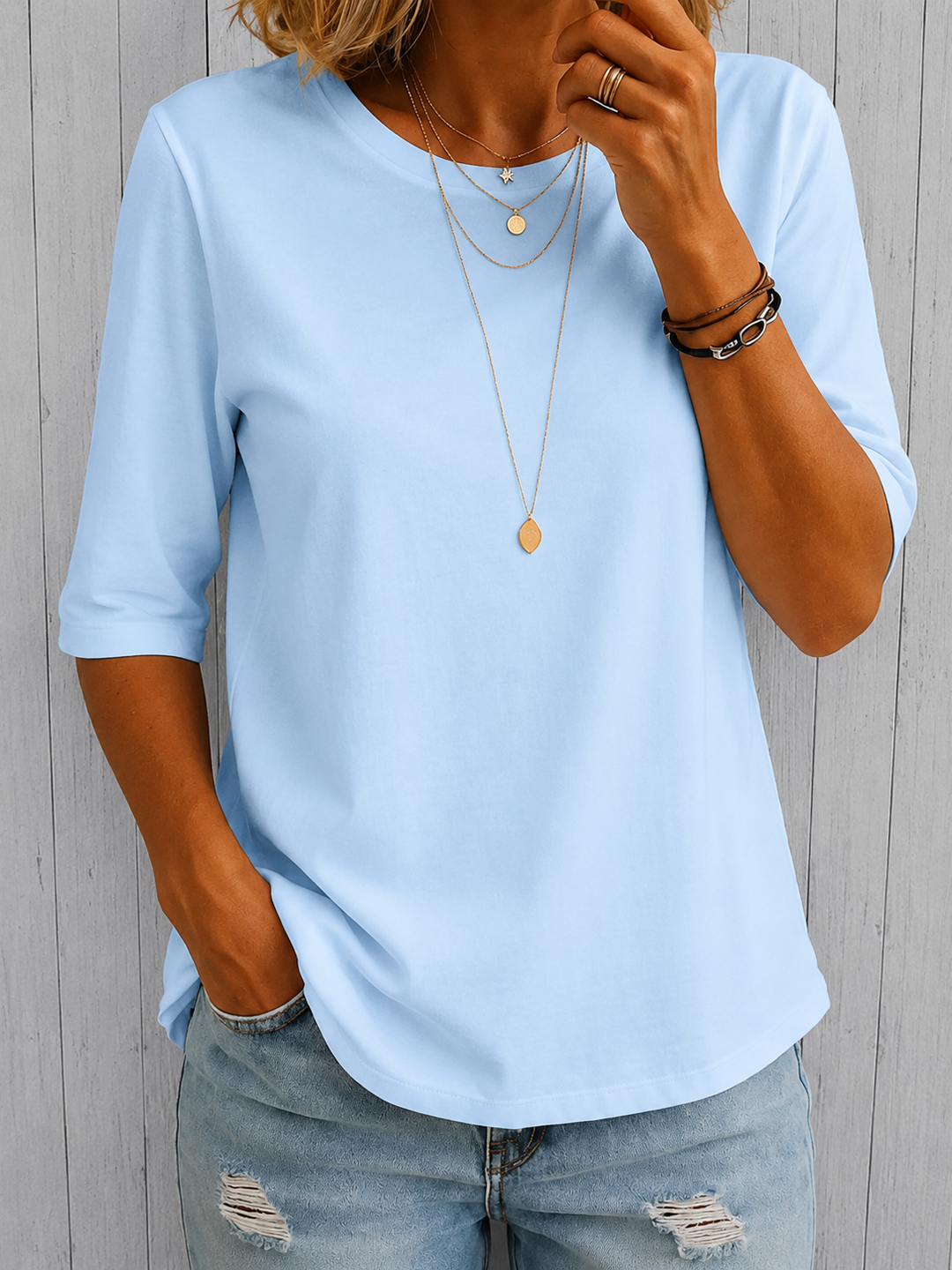 Light blue three-quarter sleeve tee with layered necklaces, worn with light blue distressed jeans, against a textured panel background.