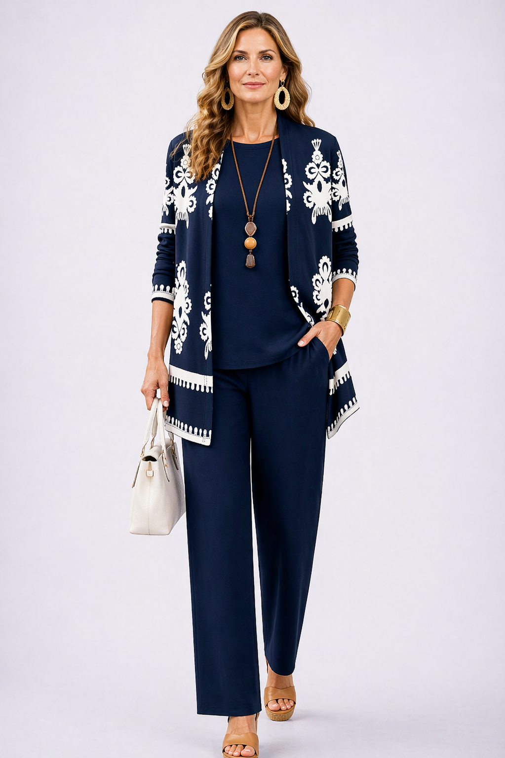 Woman wearing a navy three-piece set with elegant white patterns, posing in a studio. She carries a white handbag and wears accessories.
