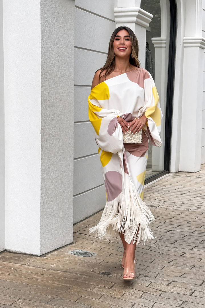 Flowing dress with yellow and mauve patterns, fringe hem, and clutch outside a white building.