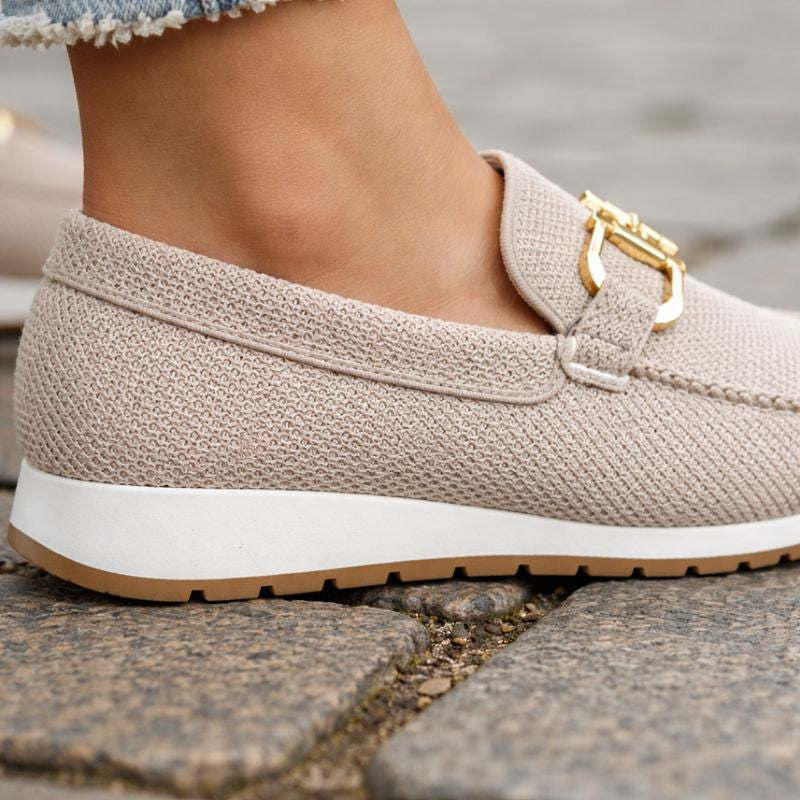Close-up of beige orthopedic loafer with gold detail on cobblestone pavement, paired with frayed jeans.