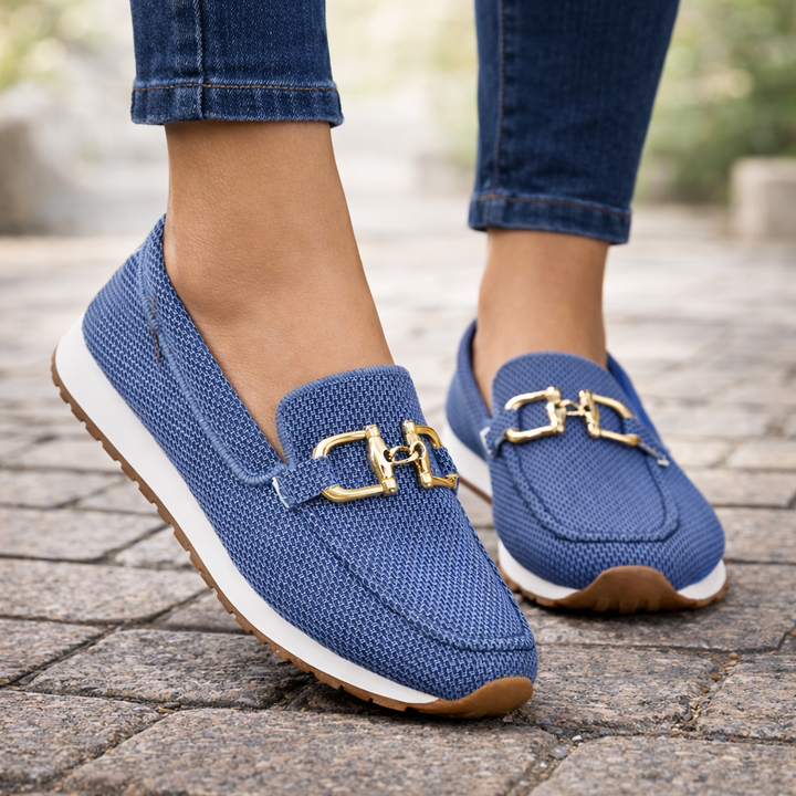 Blue orthopedic loafers with gold embellishments on cobblestone street, worn with jeans.