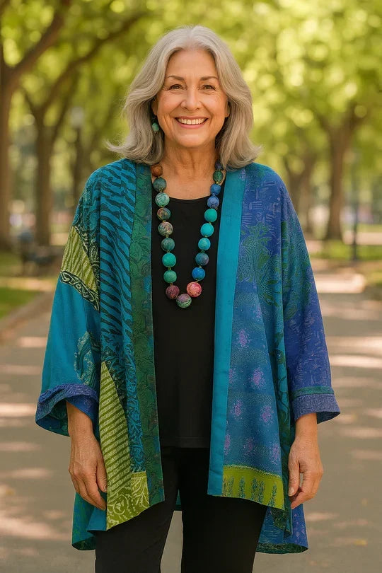 Woman wearing a colorful blue and green drape cardigan with a beaded necklace, standing outdoors on a tree-lined path.