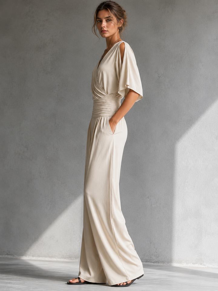 Side view of a woman wearing a cream jumpsuit with wide legs and cutout shoulders in a gray studio.
