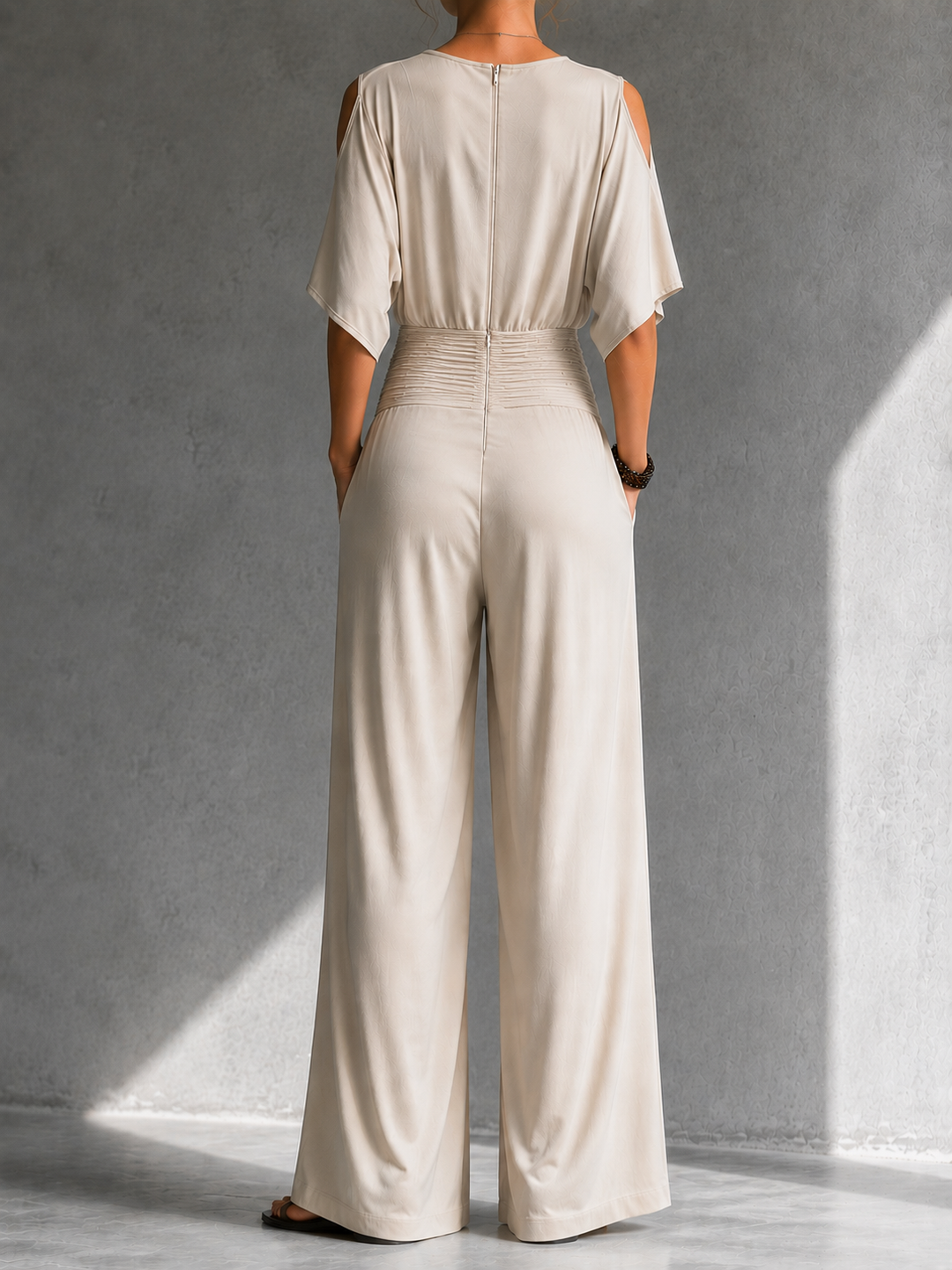 Back view of cream jumpsuit with cutout shoulders and wide legs in a softly lit gray studio.