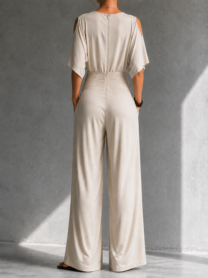 Back view of cream jumpsuit with cutout shoulders and wide legs in a softly lit gray studio.