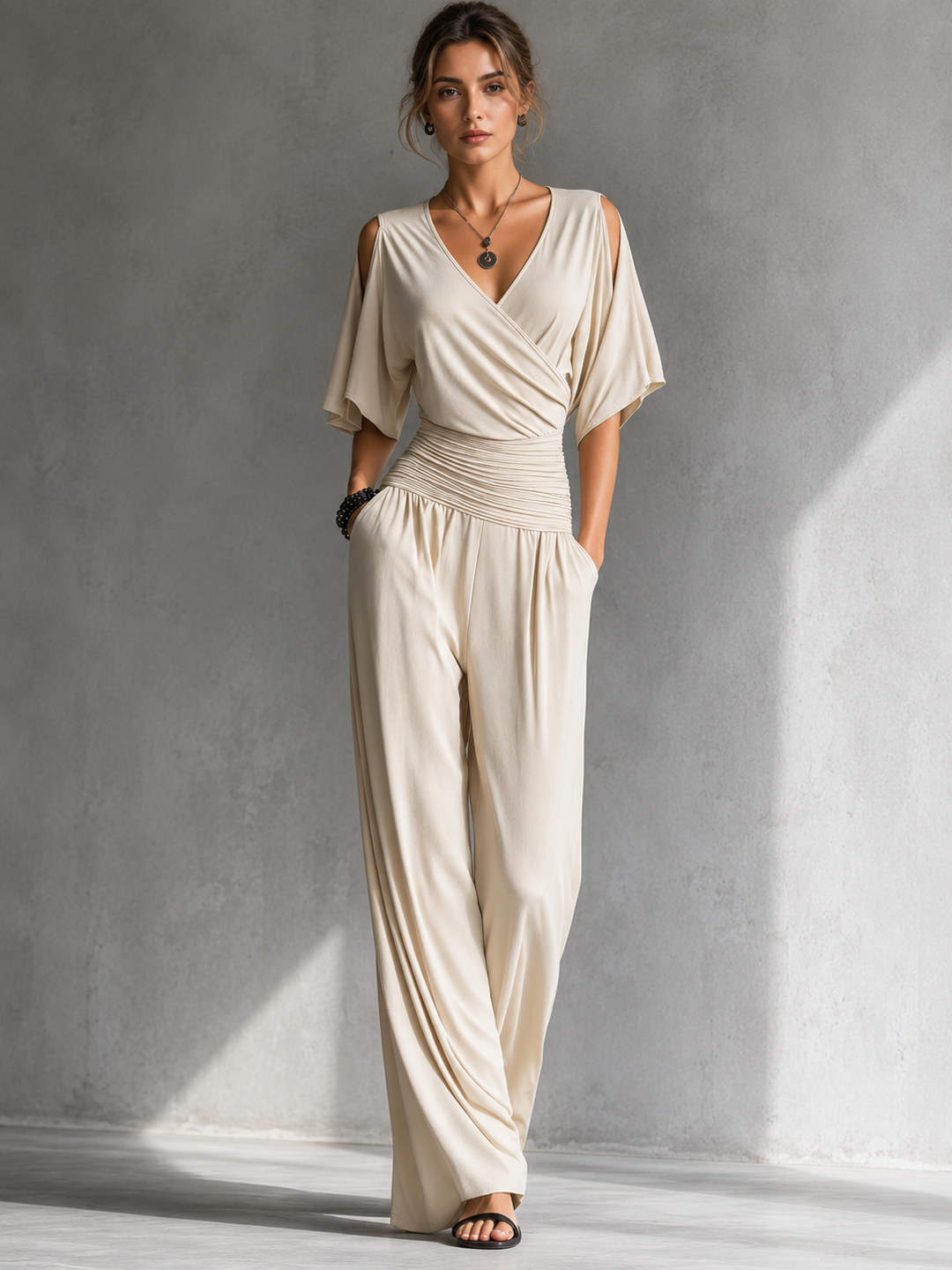 Woman wearing a cream elegant jumpsuit with cutout shoulders in a minimalistic, light gray studio setting.
