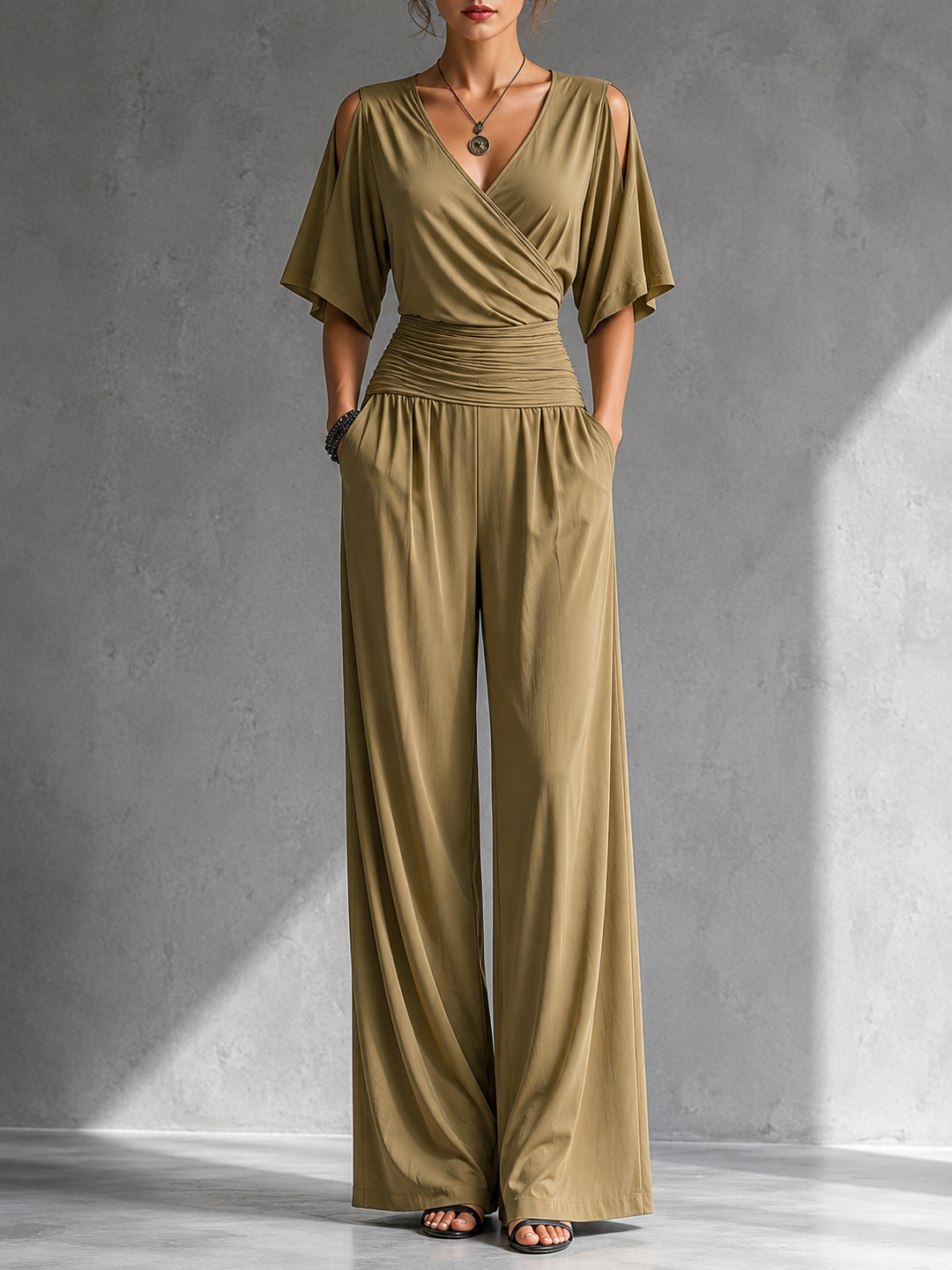 Woman in a green elegant jumpsuit with cutout shoulders and ruched waist in a simple gray studio.