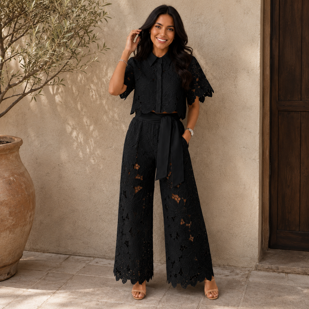 Woman in black floral lace set with wide-leg pants standing outdoors by a beige wall and wooden door.