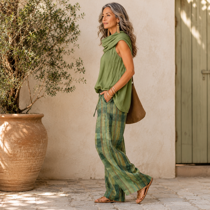 Woman in green draped top and plaid pants standing outdoors with clay pot and wooden door in the background.
