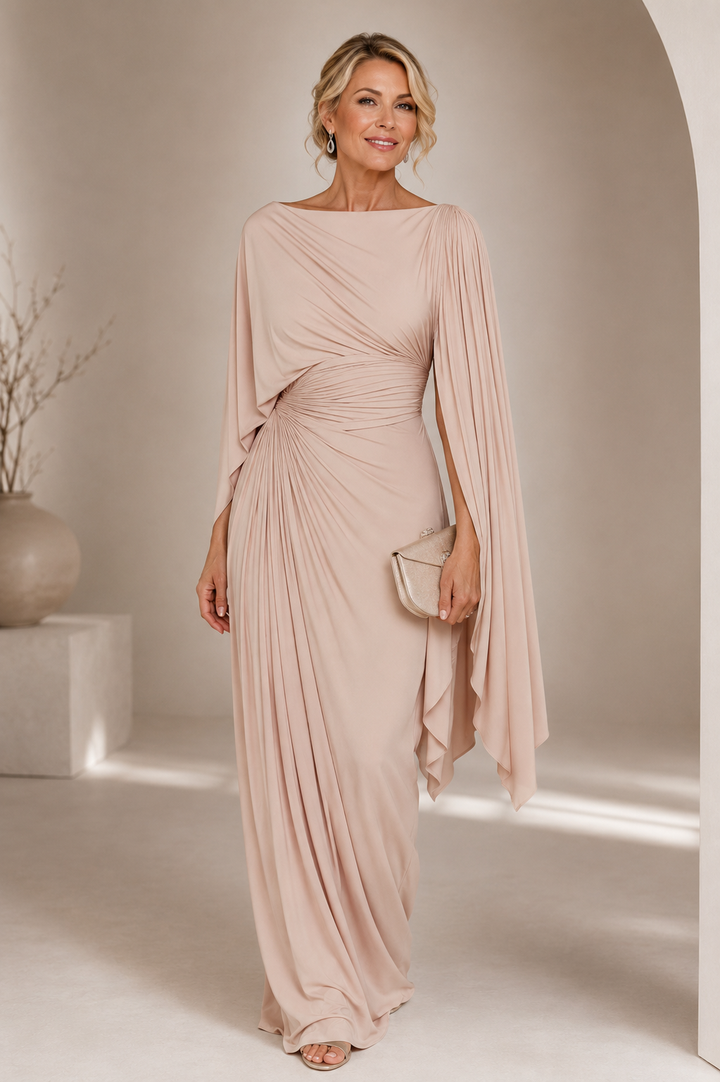 Elegant woman in a flowing blush maxi dress with draped design, holding a small clutch, standing in a minimalist indoor setting.